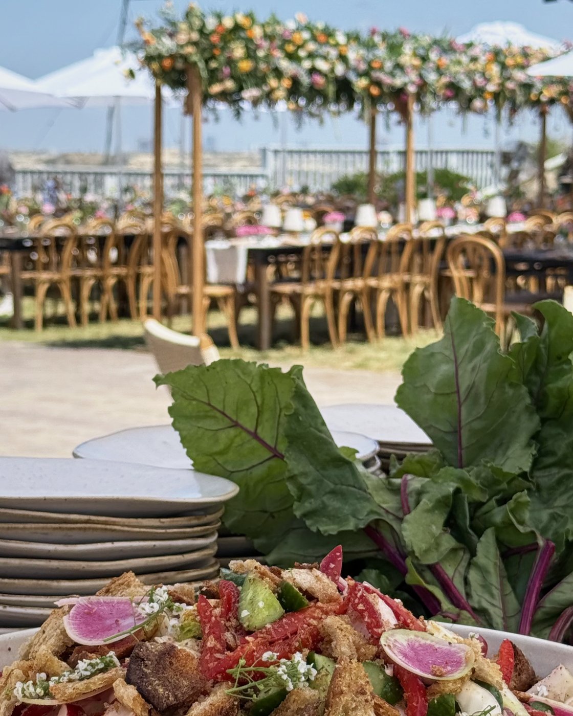 Long wedding tables under a canopy of flowers by the sea, with a seasonal salad in the foreground